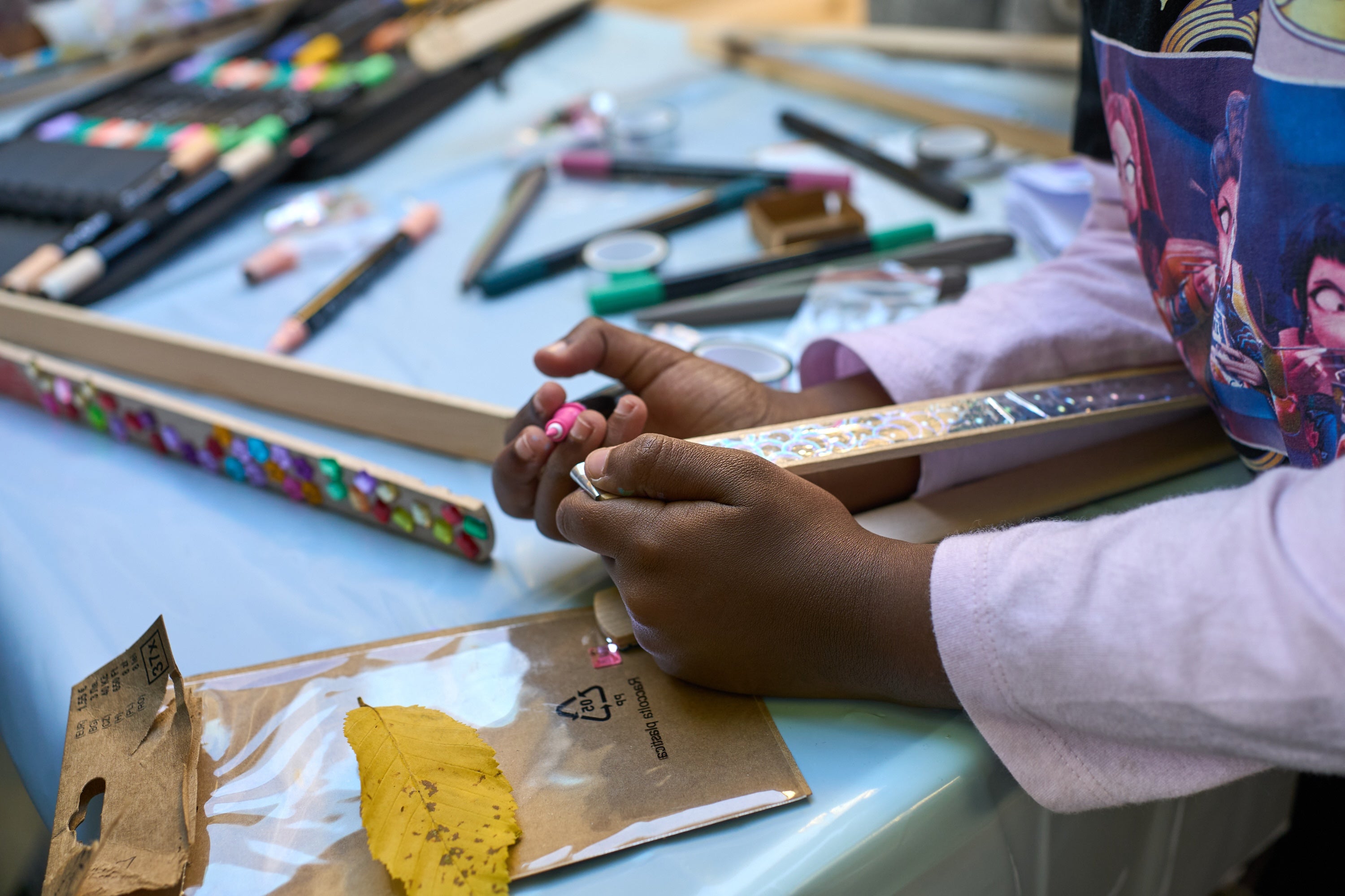 Nahaufnahme von Kinderhänden beim Basteln an einem Tisch voller Stifte, Dekosteine und Bastelmaterialien.