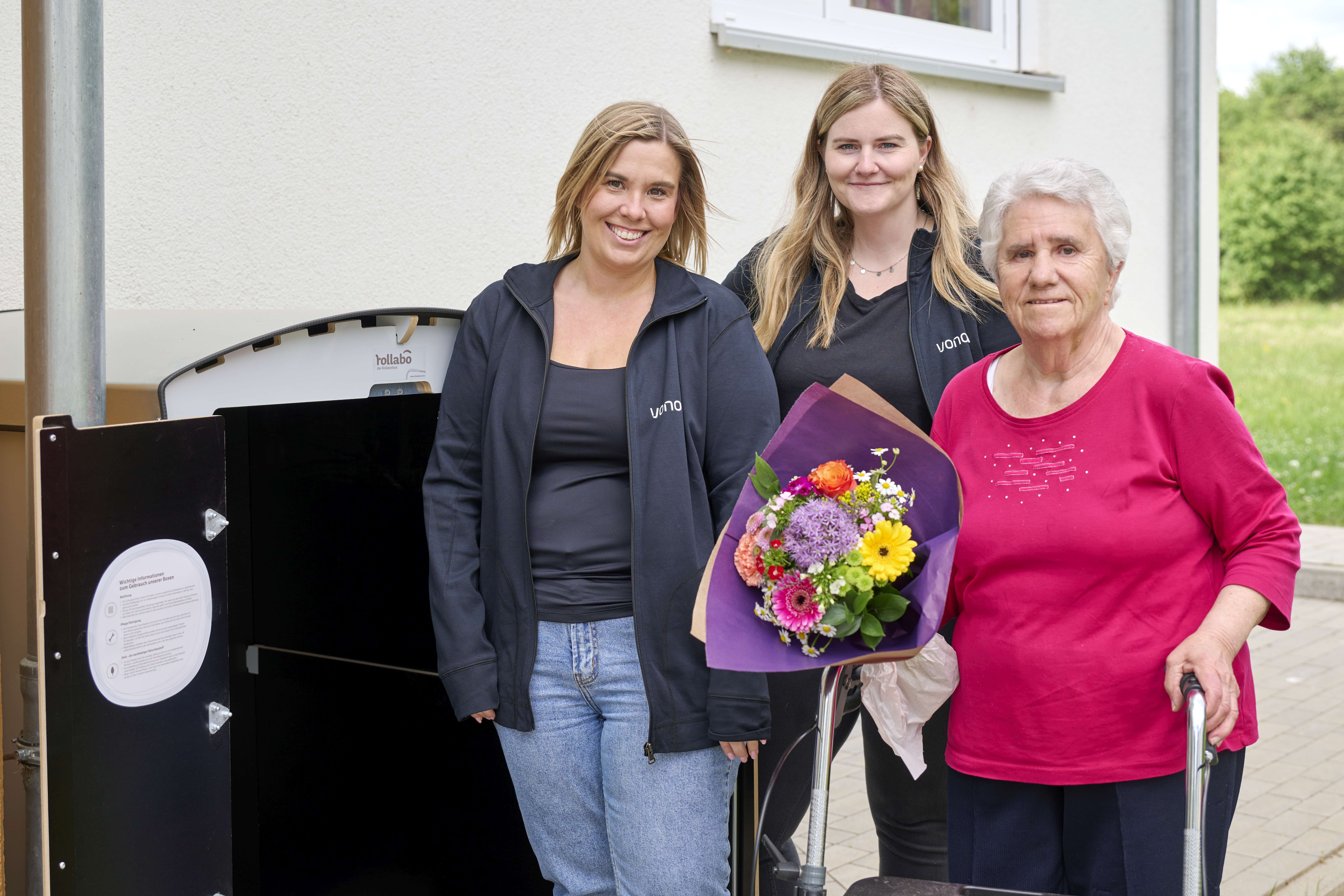 Drei Frauen, zwei mit Vonovia Jacke, die dritte mit Rollator und Blumenstrauß, stehen vor einem Wohnhaus mit einer Rollatorbox. Drei Frauen, zwei mit Vonovia Jacke, die dritte mit Rollator und Blumenstrauß, stehen vor einem Wohnhaus mit einer Rollatorbox.