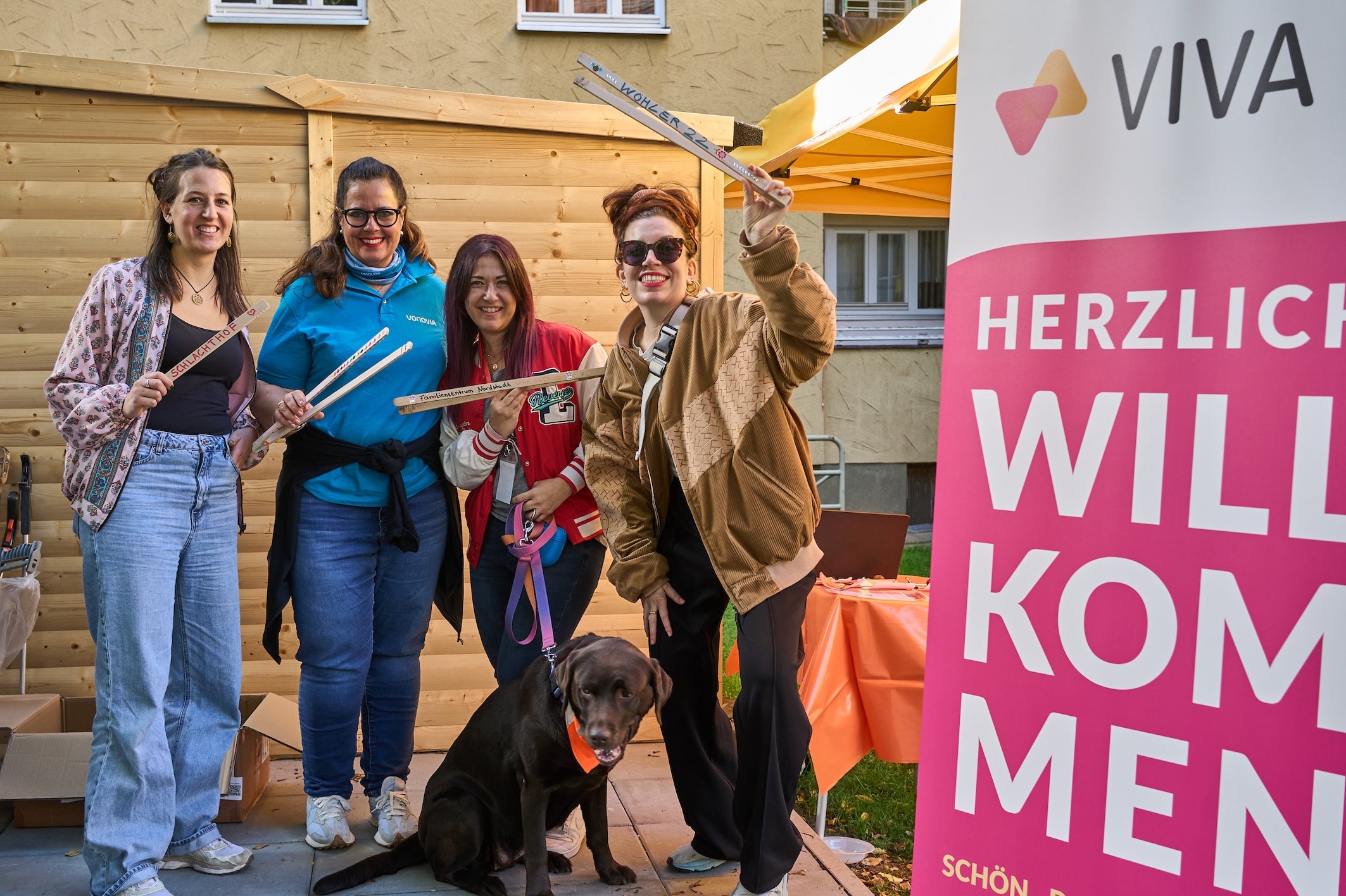 Vier lächelnde Frauen und ein brauner Hund stehen vor einem Holzschuppen bei einer Veranstaltung im Freien und halten beschriftete Holzstäbe in die Kamera; rechts ist ein rosa Aufsteller.