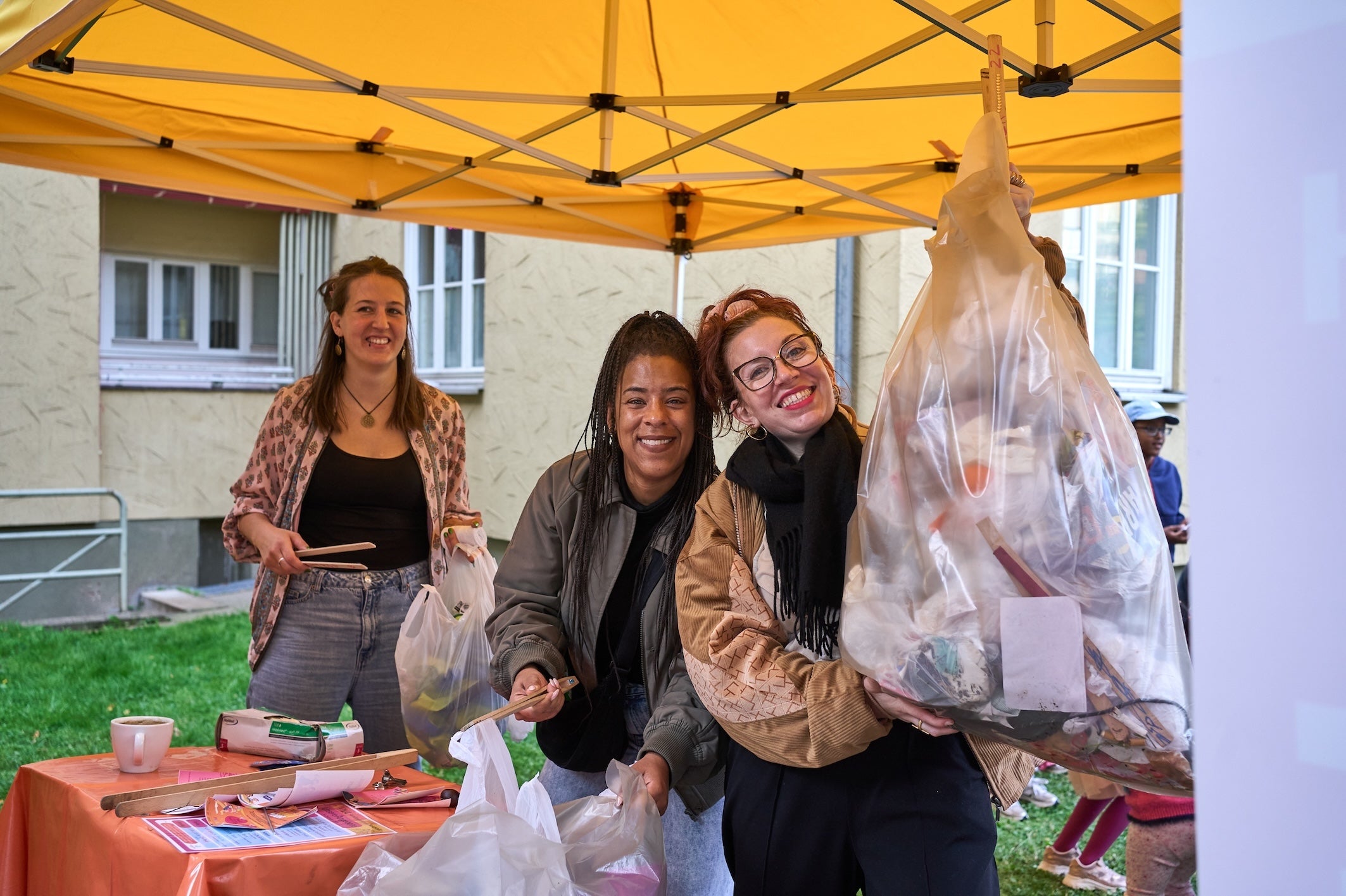 Drei lächelnde Frauen stehen unter einem gelben Pavillon bei einer Müllsammelaktion und halten gefüllte Plastikbeutel sowie Greifzangen in die Kamera.